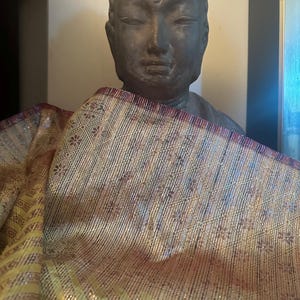 May include: A dark brown, stone Buddha statue with a gold-colored cloth draped over it. The cloth has a woven pattern of gold and brown stripes.