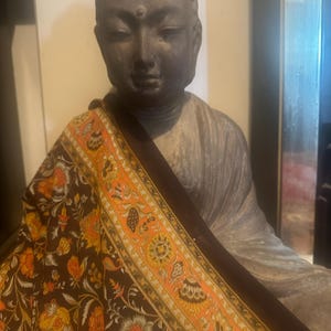 May include: A dark brown and orange patterned fabric with a floral design. The fabric is draped over a dark gray stone statue of a Buddha.