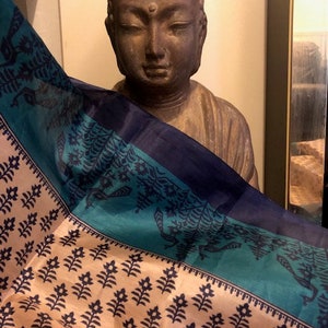 May include: A blue and teal patterned silk scarf draped over a brown statue of Buddha. The scarf has a cream background with a black floral design.