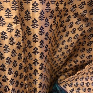 May include: A close-up of a fabric with a repeating pattern of black floral designs on a gold background. The fabric is folded in half, revealing a green fabric with a similar pattern on the bottom.