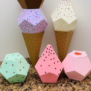 May include: Five geometric ice cream cones made of paper. The cones are in various colors and have different sprinkles. The cones are sitting on a brown countertop.