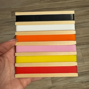 Puede incluir: Una pila de ocho bloques de madera con gomas elásticas de diferentes colores. Los colores son negro, blanco, naranja, rosa, amarillo, rojo y dos tonos más de rojo.
