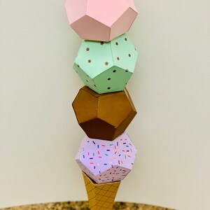 Puede incluir: Un cono de helado de papel con cuatro bolas de helado. La bola superior es rosa, la segunda es verde con lunares marrones, la tercera es marr&oacute;n y la inferior es morada con chispas de colores. El cono est&aacute; sobre una superficie marr&oacute;n.