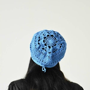 Può includere: Un berretto blu all'uncinetto con un delicato motivo floreale. Il berretto è indossato da una persona con lunghi capelli neri.