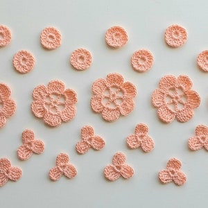 Op de afbeelding: Een verzameling van gehaakte bloemen en bladeren in perzikkleur in verschillende maten. De bloemen hebben vijf bloemblaadjes en een gat in het midden. De bladeren hebben drie lobben.