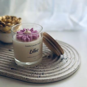 Puede incluir: Una vela blanca con una flor morada en la parte superior en un frasco de vidrio transparente. El frasco tiene una etiqueta que dice "Palette Candles Studio" y "Lilac". La vela está sentada en una bandeja texturizada gris y blanca.