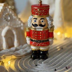 Może przedstawiać: Ozdobna figurka do dziadków do orzechów w kolorze czerwono-złotym ze szczegółową twarzą, białą peruką i czarnymi butami. Figurka ma czerwony kapelusz i kurtkę ze złotymi akcentami. Świąteczna dekoracja.