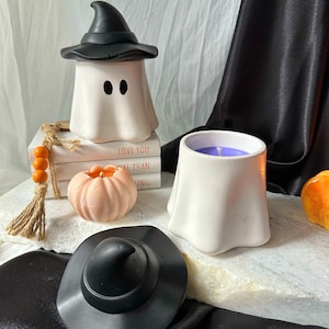 Puede incluir: Dos portavelas blancos en forma de fantasma con sombreros de bruja negros. Un portavelas tiene una vela morada dentro. Los portavelas están sobre una superficie blanca con una pila de libros, una pequeña calabaza naranja y un sombrero de bruja negro.