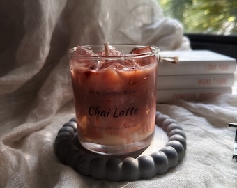 May include: A clear glass candle jar with a pink and white chai latte scented candle. The candle is on a grey beaded coaster. The label on the jar reads "Palette Candles Studio Chai Latte Crafted with love in Florida 12oz."
