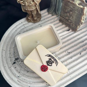 May include: A rectangular white candle with a wick sits next to a cream-colored envelope-shaped soap. The soap has a red wax seal and the words "HOGWARTS" printed on it. Decorative figurines are in the background.