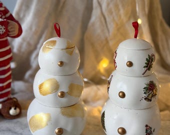 Pode incluir: Duas caixas decorativas em forma de boneco de neve. Uma é branca com pinceladas douradas e botões dourados. A outra é branca com ilustrações temáticas de Natal e botões dourados. Ambas têm laços de fita vermelha.