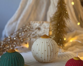 Puede incluir: Tres velas esféricas con forma de adorno navideño en verde, blanco y rojo, cada una con una parte superior dorada y pequeños detalles dorados. Las velas están sobre una bandeja circular blanca, con un árbol de Navidad dorado y otras decoraciones de fondo.