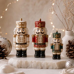 May include: Three nutcracker figurines standing in a row. The left one is white and gold, the centre one is red and gold, and the right one is green and gold. Each figure features a tall hat, a moustache, and black boots with gold trim details.