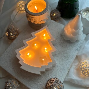 May include: A white ceramic Christmas tree candle holder with five lit tealight candles inside. The candle holder is surrounded by white fairy lights and other white ceramic Christmas decorations.