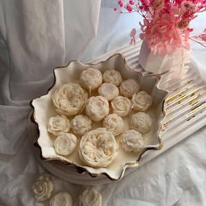 May include: A white ceramic bowl with a gold rim filled with white floral-shaped candles. The bowl is on a white surface with a small bouquet of pink flowers in a white vase.