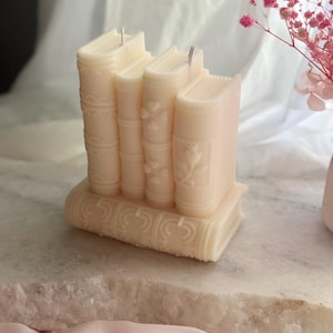 Puede incluir: Una vela blanca con forma de pila de libros con diseños en relieve detallados. La vela está sobre una superficie de mármol blanco.