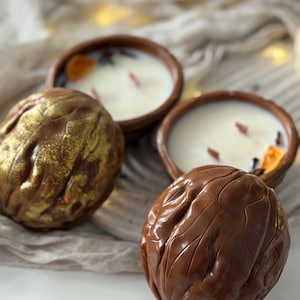May include: Close-up of two brown, walnut-shaped decorative objects and two small, round, terracotta bowls filled with white wax candles. The candles have dried orange slices and spices. One walnut is gold, the other is brown.