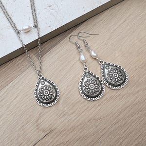 May include: A silver-toned jewelry set featuring a necklace and matching earrings. The necklace has a double chain with a pearl accent and a teardrop pendant with a floral design. The earrings also have a pearl and a teardrop pendant with a floral design.
