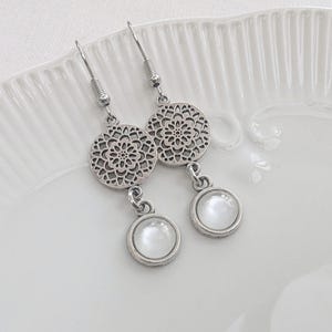 May include: A pair of silver-toned dangle earrings featuring a circular filigree charm with a cutout pattern, connected to a smaller round pendant containing a smooth, opaque white cabochon stone, suspended from standard metal earring hooks.