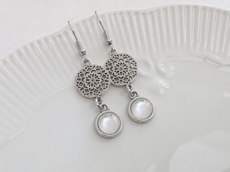 May include: A pair of silver-toned dangle earrings featuring a circular filigree charm with a cutout pattern, connected to a smaller round pendant containing a smooth, opaque white cabochon stone, suspended from standard metal earring hooks.