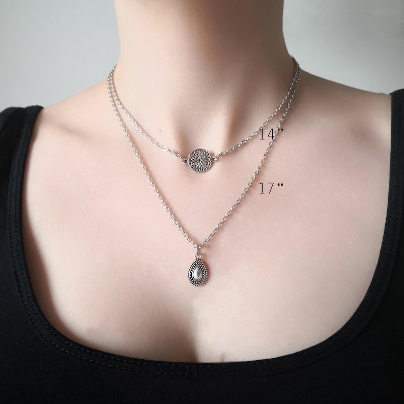 A model wearing a layered necklace with two chains. Shorter 14" chain has a antique silver plated filigree charm and the longer 17" chain has a teardrop charm.