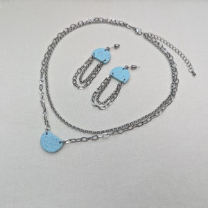 May include: A silver chain necklace with a light blue pendant and matching earrings. The pendant and earrings are shaped like half circles with a speckled texture.