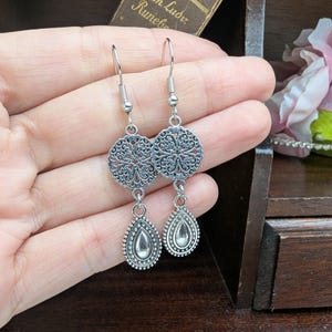 May include: A pair of silver-toned dangle earrings featuring a circular filigree charm connected to a teardrop-shaped pendant with a smooth center. The earrings are suspended from standard silver-toned metal fishhook ear wires.
