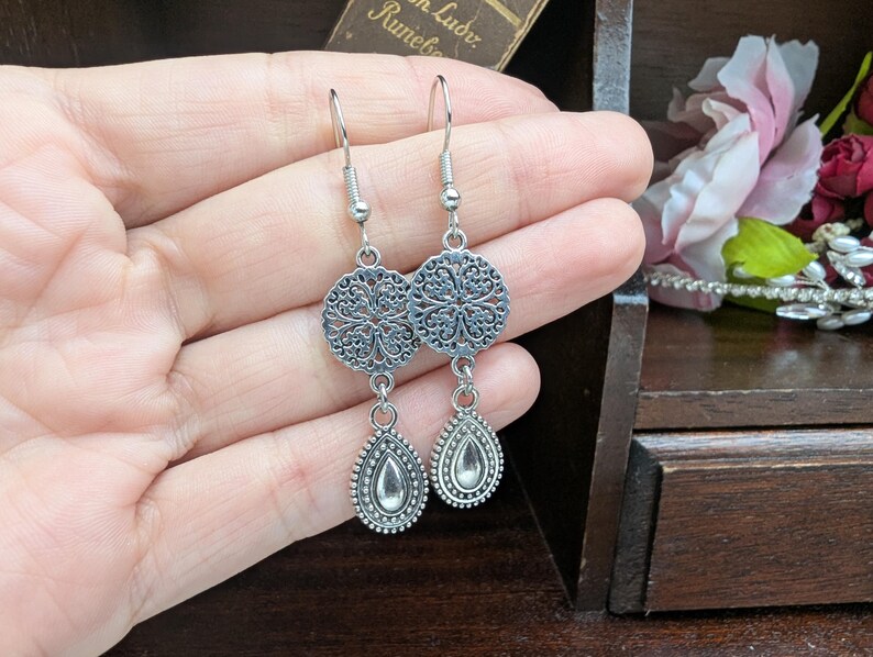 May include: A pair of silver-toned dangle earrings featuring a circular filigree charm connected to a teardrop-shaped pendant with a smooth center. The earrings are suspended from standard silver-toned metal fishhook ear wires.