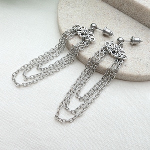 May include: Silver-toned dangle earrings. Each earring features a floral design at the stud and three strands of chain. The earrings are displayed on a light-colored, textured surface.