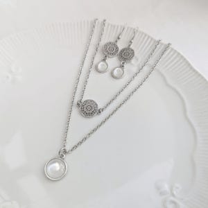 May include: A silver-toned jewelry set consisting of a double-layered chain necklace and a pair of matching drop earrings. The necklace features a circular pendant with a white cabochon and a circular filigree charm. The earrings feature matching filigree discs and white cabochons.