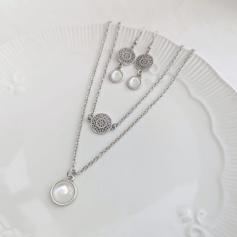 May include: A silver-toned jewelry set consisting of a double-layered chain necklace and a pair of matching drop earrings. The necklace features a circular pendant with a white cabochon and a circular filigree charm. The earrings feature matching filigree discs and white cabochons.