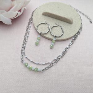 May include: A silver chain necklace with a double layer of chains and green crystal beads. The necklace is paired with a set of silver hoop earrings with green crystal beads dangling from each hoop.