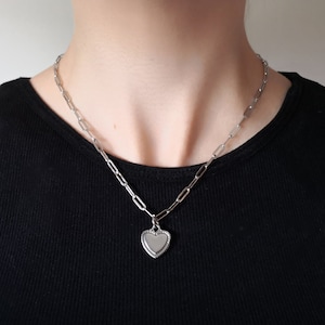 May include: A silver-toned necklace featuring a chain of elongated oval links and a central heart-shaped pendant. The pendant has a smooth, reflective center framed by a textured, beaded border, suspended from the chain by a small jump ring.