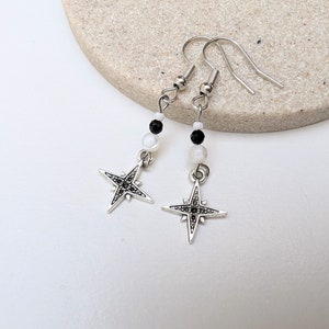 May include: A pair of silver star earrings with black and white beads. The stars have a detailed design and are accented with small black rhinestones.