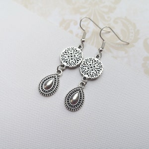 May include: A pair of silver-toned dangle earrings featuring a circular filigree charm connected to a teardrop-shaped pendant with a smooth center and beaded border. The earrings are attached to standard silver-toned metal ear wires.