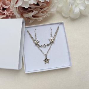 May include: A silver-toned jewelry set consisting of a multi-layered chain necklace and a pair of matching drop earrings. The necklace and earrings feature textured star-shaped charms and small open star accents, all presented in a white box.