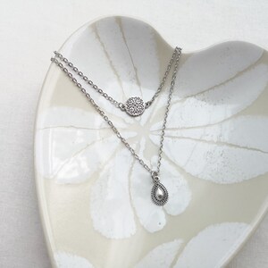 May include: A silver-toned double-layered necklace featuring a small circular pendant with an intricate filigree pattern on the upper chain and a teardrop-shaped charm with a beaded border on the lower chain, crafted from polished metal links.