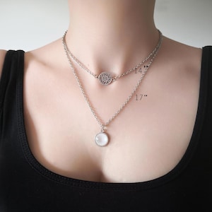 May include: A silver-toned layered necklace featuring two chains. The shorter 14-inch chain has a circular filigree charm, while the longer 17-inch chain holds a round, smooth white cabochon pendant. Both chains are delicate and metallic.