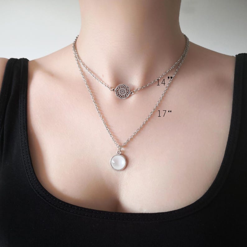 May include: A silver-toned layered necklace featuring two chains. The shorter 14-inch chain has a circular filigree charm, while the longer 17-inch chain holds a round, smooth white cabochon pendant. Both chains are delicate and metallic.