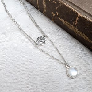 May include: A silver-toned double-layered chain necklace featuring a circular filigree charm with an intricate openwork pattern and a second pendant consisting of a smooth, round, iridescent white cabochon stone set in a simple silver-toned metal bezel.