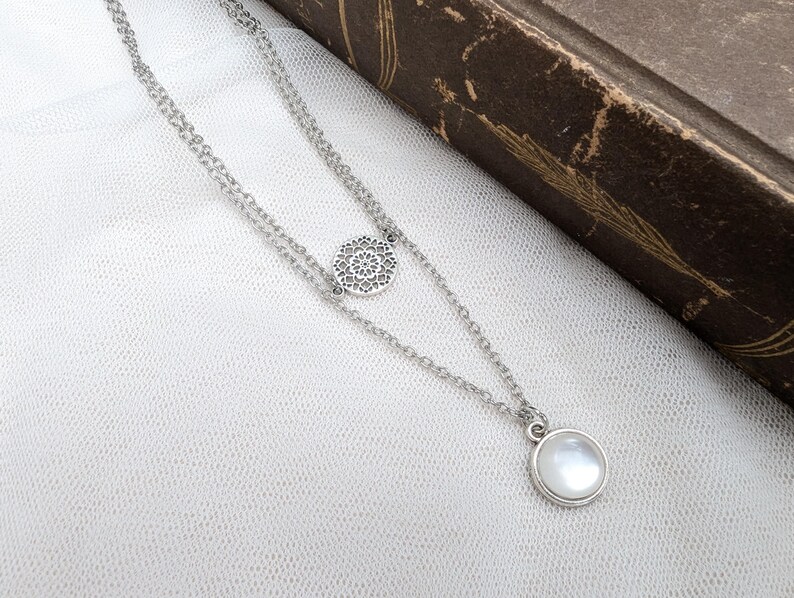 May include: A silver-toned double-layered chain necklace featuring a circular filigree charm with an intricate openwork pattern and a second pendant consisting of a smooth, round, iridescent white cabochon stone set in a simple silver-toned metal bezel.