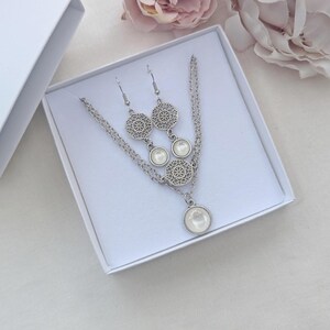 May include: A silver-toned jewelry set consisting of a necklace and a pair of drop earrings. Each piece features a circular filigree metal charm paired with a smooth, round, white cabochon stone, presented in a white gift box.