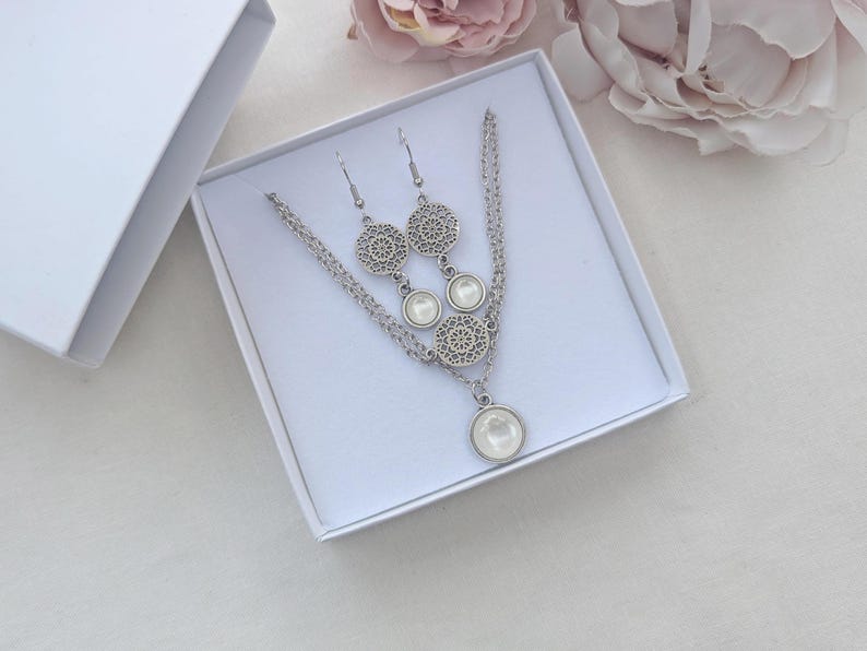 May include: A silver-toned jewelry set consisting of a necklace and a pair of drop earrings. Each piece features a circular filigree metal charm paired with a smooth, round, white cabochon stone, presented in a white gift box.