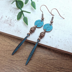 May include: A pair of copper-toned earrings with a turquoise finish. Each earring has a turquoise oval with a detailed pattern, a copper bead, and a long, pointed turquoise dangle. The earrings are displayed on a wooden surface.