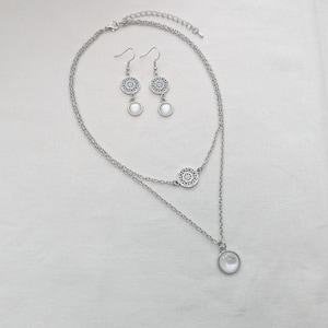 May include: A silver-toned layered necklace and matching drop earrings set. The necklace features two delicate chains, a circular filigree charm, and a round white cabochon pendant. The earrings mirror the pendant design with filigree tops.
