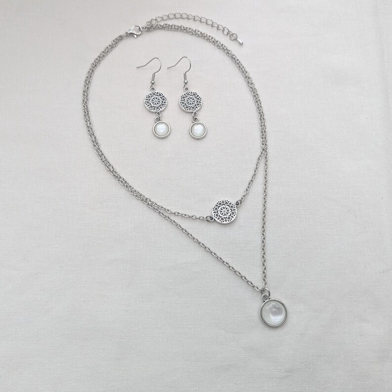 May include: A silver-toned layered necklace and matching drop earrings set. The necklace features two delicate chains, a circular filigree charm, and a round white cabochon pendant. The earrings mirror the pendant design with filigree tops.