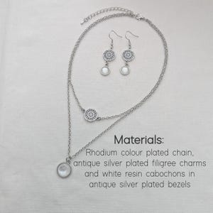 May include: A double-layered silver-toned necklace and matching drop earrings. The necklace features a filigree charm and a white resin cabochon pendant. Text reads: Materials: Rhodium colour plated chain, antique silver plated filigree charms and white resin cabochons in antique silver plated bezels.