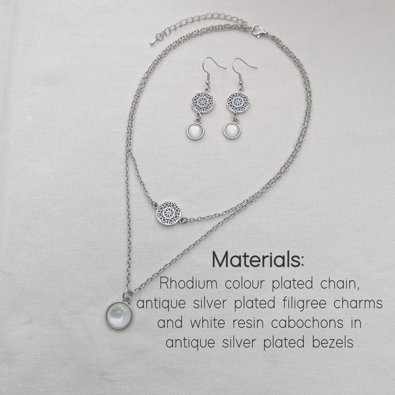 May include: A double-layered silver-toned necklace and matching drop earrings. The necklace features a filigree charm and a white resin cabochon pendant. Text reads: Materials: Rhodium colour plated chain, antique silver plated filigree charms and white resin cabochons in antique silver plated bezels.