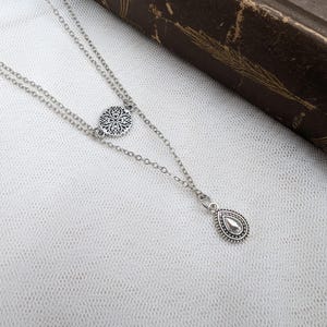 May include: A silver-toned double-layered chain necklace featuring a small circular charm with an intricate filigree pattern and a teardrop-shaped pendant with a beaded border. The metal components have an oxidized finish to highlight the details.