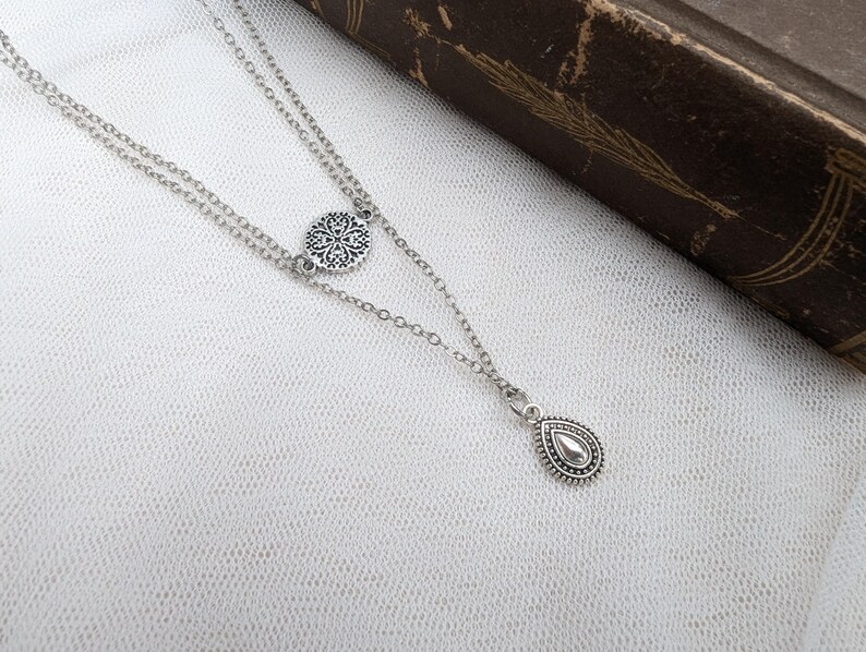 May include: A silver-toned double-layered chain necklace featuring a small circular charm with an intricate filigree pattern and a teardrop-shaped pendant with a beaded border. The metal components have an oxidized finish to highlight the details.